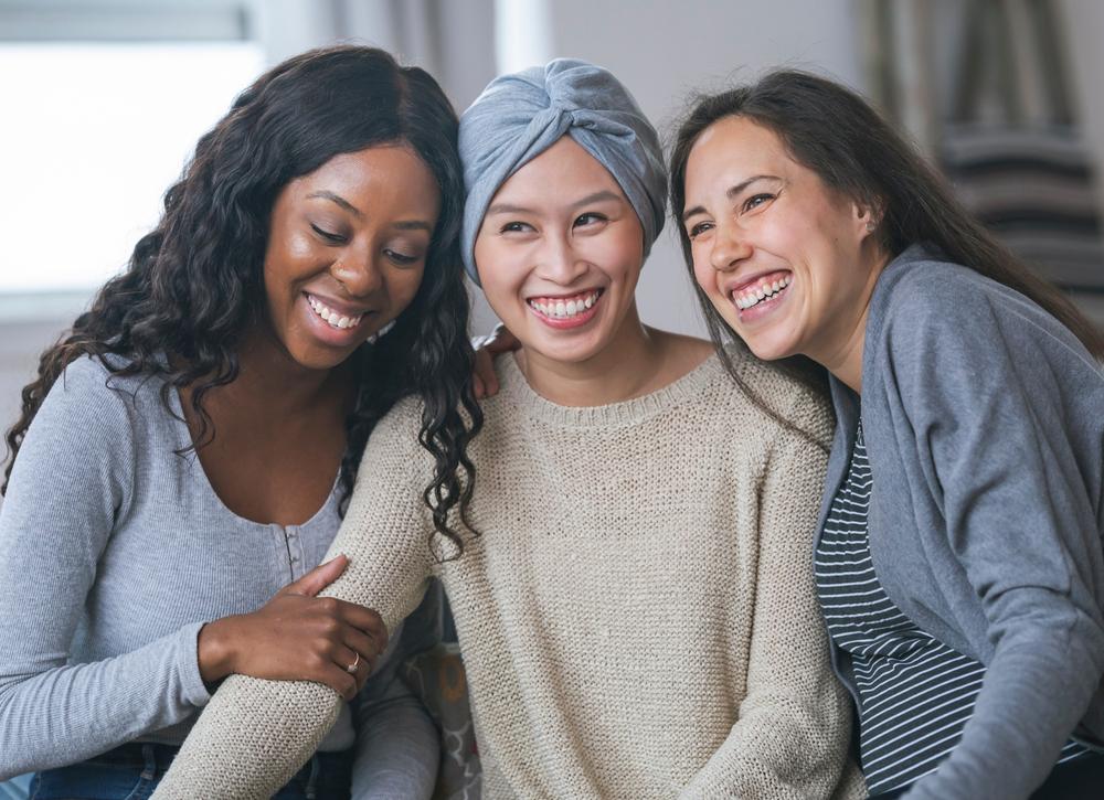 Un trio de femmes souriantes, amies, dont l'une porte les stigmates de la maladie du cancer du sein, ainsi qu'un foulard